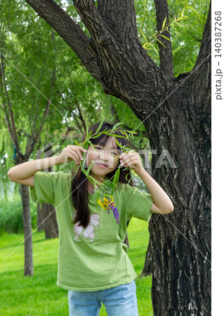 A playful young girl hiding behind a willow branch garland in front of a massive tree trunk. A playful young girl hiding behind a willow branch garland in front of a massive tree trunk. 140387268