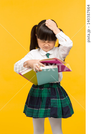 A confused schoolgirl scratching her head while holding textbooks against a yellow background. 140387664