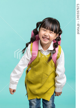 A cheerful Asian female student, with her hair in gourd braids and carrying a pink backpack, stands against a blue-green background with a back-to-school theme. A cheerful Asian female student, with her hair in gourd braids and carrying a pink backpack, stands against a blue-green background with a back-to-school theme. 140387673