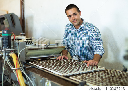 Farmer man working with automatic filling and seeding line for growing containerized seedlings of decorative plants in greenhouse 140387948