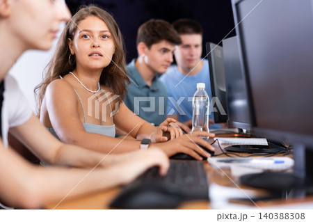 Positive young girl student sits at computer during lesson and asks question to neighbor classmate Positive young girl student sits at computer during lesson and asks question to neighbor classmate 140388308