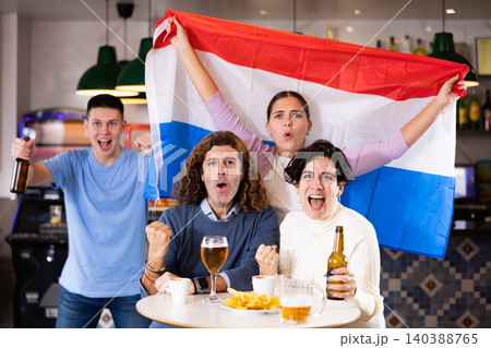 Company of excited young adult sports fans waving Dutch flag and supporting national team with beer in pub Company of excited young adult sports fans waving Dutch flag and supporting national team with beer in pub 140388765