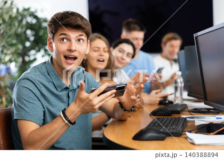 At computer class, young guy student sits with mobile phone and listens admiringly to teacher At computer class, young guy student sits with mobile phone and listens admiringly to teacher 140388894