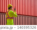 Asian man worker talking on walkie talkie for control loading containers box while standing in front of red containers at cargo shipping yard 140395326