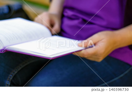 Book reading in hands of a woman wearing purple t-shirt. Close up of female student holding open novel or textbook, studying, self education and literature concept with selective focus Book reading in hands of a woman wearing purple t-shirt. Close up of female student holding open novel or textbook, studying, self education and literature concept with selective focus 140396730