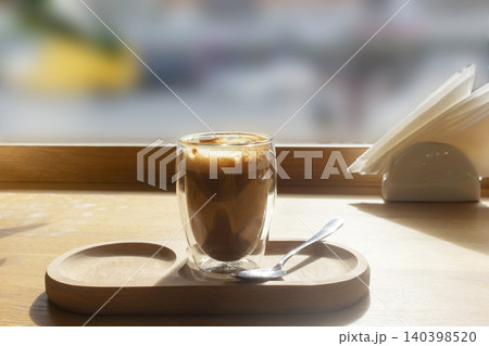 Close up photo of a glass cup of coffee with foam on a wooden tray, accompanied by a spoon and napkin. 140398520