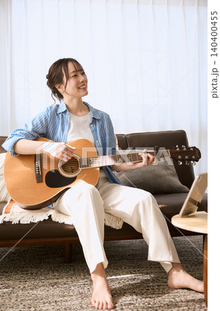 リビングのソファでアコースティックギターを弾く笑顔の若い女性 リビングのソファでアコースティックギターを弾く笑顔の若い女性 140400455
