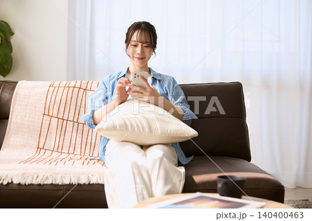 リビングのソファでスマートフォンを操作する笑顔の若い女性 140400463
