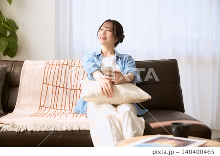 リビングのソファでスマホを持ちくつろぐ笑顔の若い女性 リビングのソファでスマホを持ちくつろぐ笑顔の若い女性 140400465