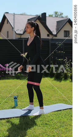 Vertical video. Attractive woman trains her buttocks and leg muscles on a blue carpet outdoors, on the grass in nature. Sports female doing outdoor training. Vertical video. Attractive woman trains her buttocks and leg muscles on a blue carpet outdoors, on the grass in nature. Sports female doing outdoor training. 140400866