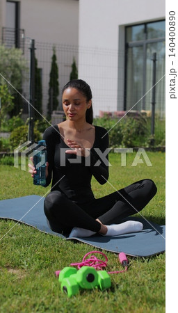Vertical video. Young sport woman resting, drink water after workout in backyard. Sports female doing outdoor training. 140400890