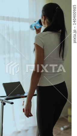 Vertical video. Athletic woman in sportswear running and drink water on treadmill during home workout. Back view. Vertical video. Athletic woman in sportswear running and drink water on treadmill during home workout. Back view. 140400985