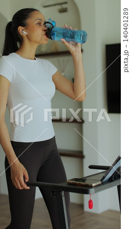 Vertical video. Athletic woman in sportswear running and drink water on treadmill during home workout. Vertical video. Athletic woman in sportswear running and drink water on treadmill during home workout. 140401289