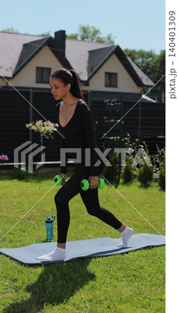 Vertical video. Athletic young woman in sportswear training on the lawn of the house, performing squats with dumbbells in her hands, enjoying a healthy lifestyle. Athletic woman doing training Vertical video. Athletic young woman in sportswear training on the lawn of the house, performing squats with dumbbells in her hands, enjoying a healthy lifestyle. Athletic woman doing training 140401309