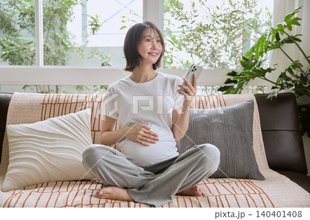 ソファでスマートフォンを持ちお腹に手を当てる妊婦 140401408