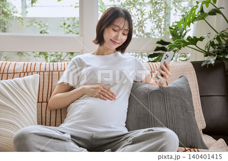 ソファでスマートフォンを見ながらお腹に手を当てる妊婦 ソファでスマートフォンを見ながらお腹に手を当てる妊婦 140401415