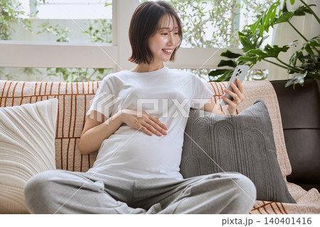 ソファでスマートフォンを見ながらお腹に手を当てる妊婦 ソファでスマートフォンを見ながらお腹に手を当てる妊婦 140401416