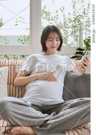 ソファでスマホを見ながらお腹に手を当てる笑顔の妊婦 140401419