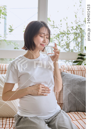 ソファに座り水を飲みながらお腹を押さえる妊婦 ソファに座り水を飲みながらお腹を押さえる妊婦 140401428