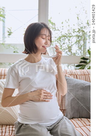 ソファに座り水を飲みながらお腹を押さえる妊婦 ソファに座り水を飲みながらお腹を押さえる妊婦 140401429