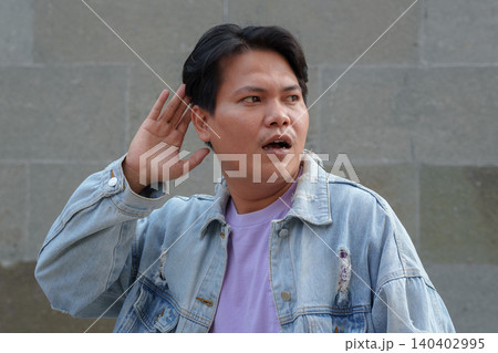 Surprised Young Asian Man Cupping Hand to Ear Listening Carefully Against Stone Wall Surprised Young Asian Man Cupping Hand to Ear Listening Carefully Against Stone Wall 140402995