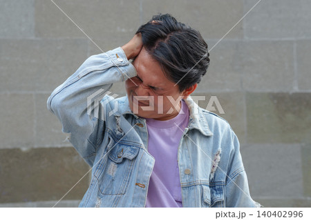 Stressed Young Asian Man Holding His Head with Painful Expression Against Stone Wall Stressed Young Asian Man Holding His Head with Painful Expression Against Stone Wall 140402996