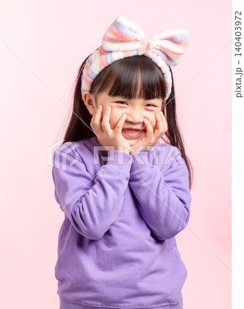 A close-up portrait of a happy Asian girl with bangs, wearing a striped headband and a purple sweatshirt, smiling brightly while cupping her cheeks in a cute, happy pose against a light pink A close-up portrait of a happy Asian girl with bangs, wearing a striped headband and a purple sweatshirt, smiling brightly while cupping her cheeks in a cute, happy pose against a light pink 140403972