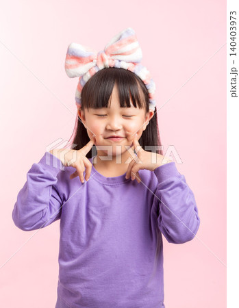 A playful and bashful studio portrait of an Asian elementary girl with bangs and a striped headband, wearing a purple sweatshirt, shyly pointing at her cheeks with her index fingers and smiling A playful and bashful studio portrait of an Asian elementary girl with bangs and a striped headband, wearing a purple sweatshirt, shyly pointing at her cheeks with her index fingers and smiling 140403973