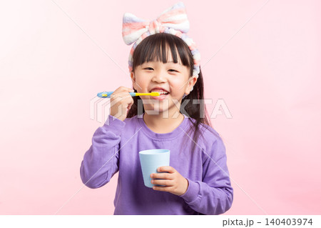 A young Asian elementary girl with long dark hair, bangs, and a striped headband, wearing a purple sweatshirt, happily brushing her teeth while holding a blue cup against a solid light pink studio A young Asian elementary girl with long dark hair, bangs, and a striped headband, wearing a purple sweatshirt, happily brushing her teeth while holding a blue cup against a solid light pink studio 140403974