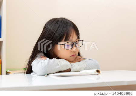 Tired or bored little girl wearing glasses and leaning on her arms over a book at a desk. Tired or bored little girl wearing glasses and leaning on her arms over a book at a desk. 140403996