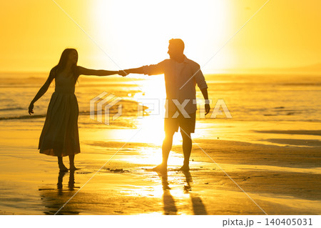 Couple walks hand in hand on beach during sunset with bright sun reflecting on water 140405031