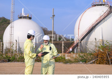 Two workers inspect storage tanks. They wear protective suits and helmets, holding tools. Two workers inspect storage tanks. They wear protective suits and helmets, holding tools. 140405955