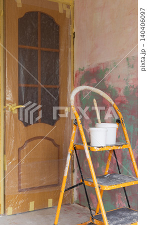 Home renovation and interior painting project with yellow stepladder, paint buckets, and brush in room under refurbishment. Wooden door protected with plastic sheeting and masking tape Home renovation and interior painting project with yellow stepladder, paint buckets, and brush in room under refurbishment. Wooden door protected with plastic sheeting and masking tape 140406097