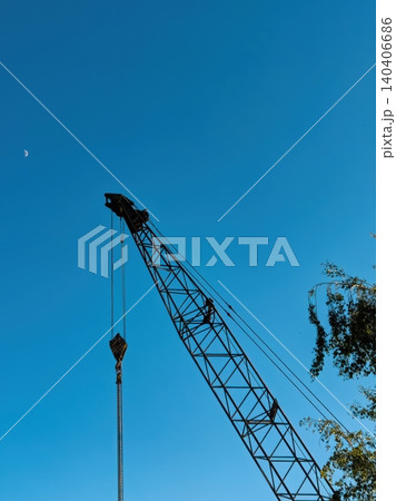 Industrial crane against clear blue sky with moon and tree foliage in the background Industrial crane against clear blue sky with moon and tree foliage in the background 140406686