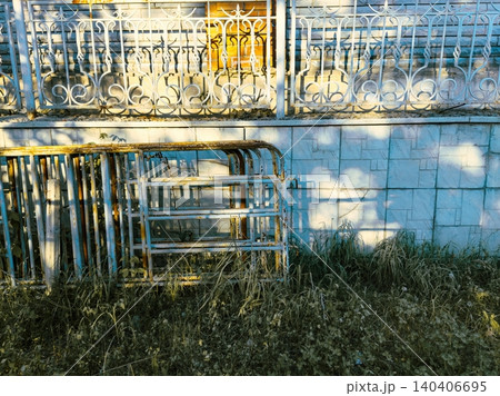 Rusty metal bed frames leaning against weathered wall in overgrown grassy yard with shadow patterns 140406695