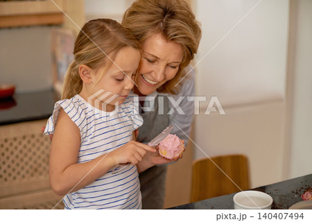 Woman, child and icing cupcake in kitchen for baking decoration with grandmother, bonding or ingredients. Female person, girl and recipe for sweet dessert at counter or teaching, helping or support 140407494
