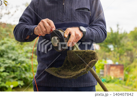 Man changing orange cutting line for string trimmer spool. Hands of gardener repairing lawn mower equipment for cutting grass. DIY garden tool maintenance and backyard landscaping work. Man changing orange cutting line for string trimmer spool. Hands of gardener repairing lawn mower equipment for cutting grass. DIY garden tool maintenance and backyard landscaping work. 140407842