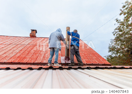 Two men worker installing metal chimney on red house roof. Professional roofers doing repair of smoke ventilation system. Maintenance service and external building construction work. Two men worker installing metal chimney on red house roof. Professional roofers doing repair of smoke ventilation system. Maintenance service and external building construction work. 140407843