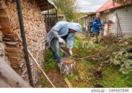 Two men working in courtyard to clear cut rowan branches. Seasonal garden maintenance and tree pruning. Helpers organizing wood and gathering garden waste in backyard for cleanup. Two men working in courtyard to clear cut rowan branches. Seasonal garden maintenance and tree pruning. Helpers organizing wood and gathering garden waste in backyard for cleanup. 140407846