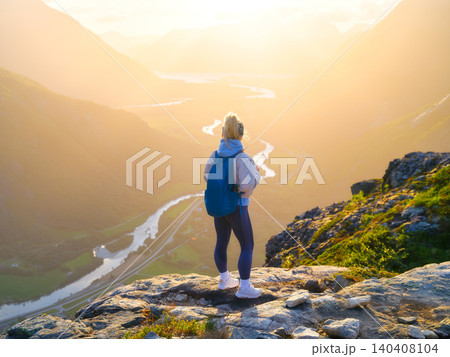 Athletic girl with a hiking backpack stands on a rock overlooking a sun-drenched mountain valley Athletic girl with a hiking backpack stands on a rock overlooking a sun-drenched mountain valley 140408104