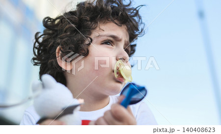 Young boy with curly hair using pacifier and holding toy outdoors 140408623
