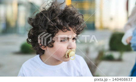 Young boy with curly hair thinking outdoors, having a pacifier in mouth Young boy with curly hair thinking outdoors, having a pacifier in mouth 140408637