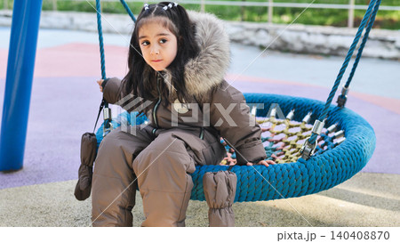 Little girl playing on a blue nest swing at a colorful playground in winter Little girl playing on a blue nest swing at a colorful playground in winter 140408870