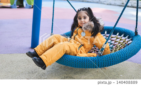 Young girl in winter clothes happily swinging on a nest swing at a colorful playground 140408889