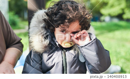 Young boy crying and rubbing his eye, holding a pacifier while feeling upset outdoors Young boy crying and rubbing his eye, holding a pacifier while feeling upset outdoors 140408913