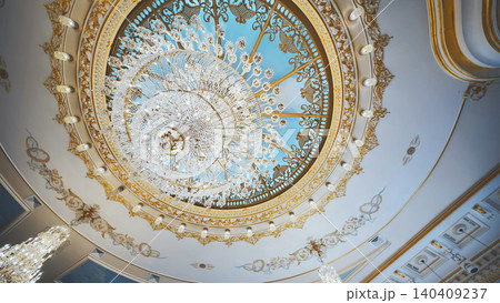 Low angle view of a grand crystal chandelier in a luxury hall. Ornate gold dome with painted blue sky. Low angle view of a grand crystal chandelier in a luxury hall. Ornate gold dome with painted blue sky. 140409237