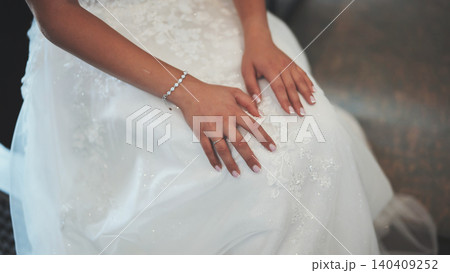 Bride sitting on chair wearing elegant white wedding dress with floral embroidery, getting ready for wedding ceremony 140409252