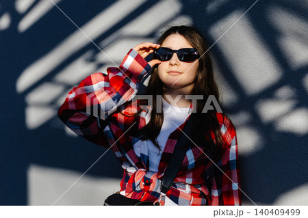 Young student adjusting sunglasses while leaning against wall with shadows 140409499