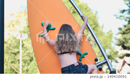 Young girl climbing an orange playground wall, reaching for the next green rock hold Young girl climbing an orange playground wall, reaching for the next green rock hold 140410091