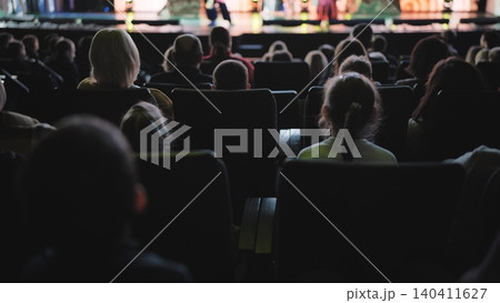 People sitting in rows, enjoying a show. Audience views the stage in a dim auditorium People sitting in rows, enjoying a show. Audience views the stage in a dim auditorium 140411627